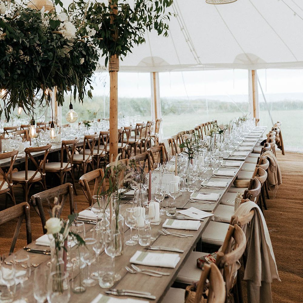 Marquee wedding reception complete with wooden banquet tables, green foliage installations hanging above and wicker lights