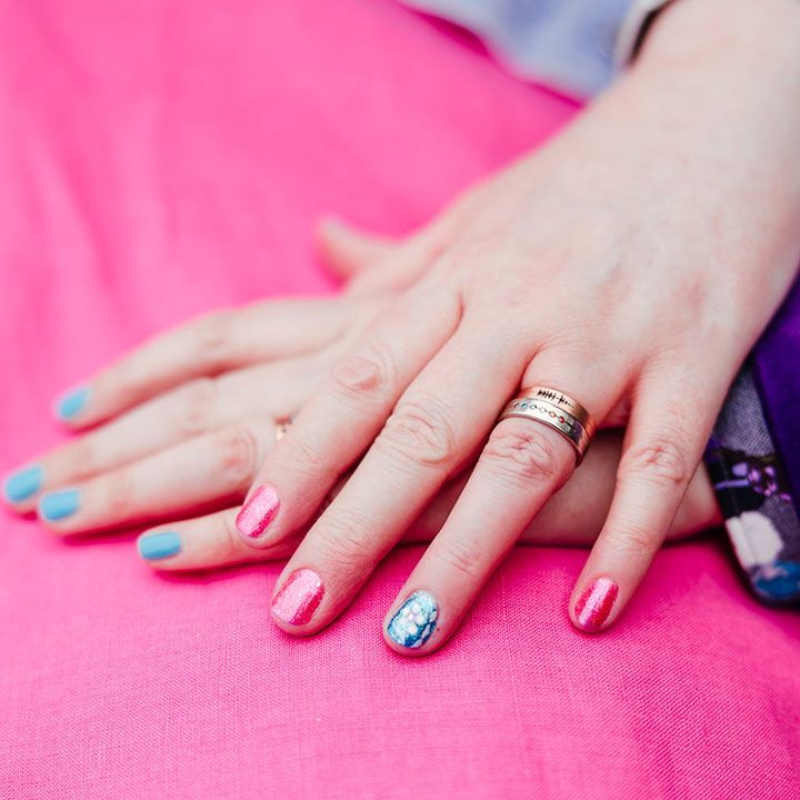 Brides with complimenting sparkly pink and blue nail varnish showing off their wedding rings in each other's hands on top of pink wedding dress with the sleeve of a purple velvet blazer 