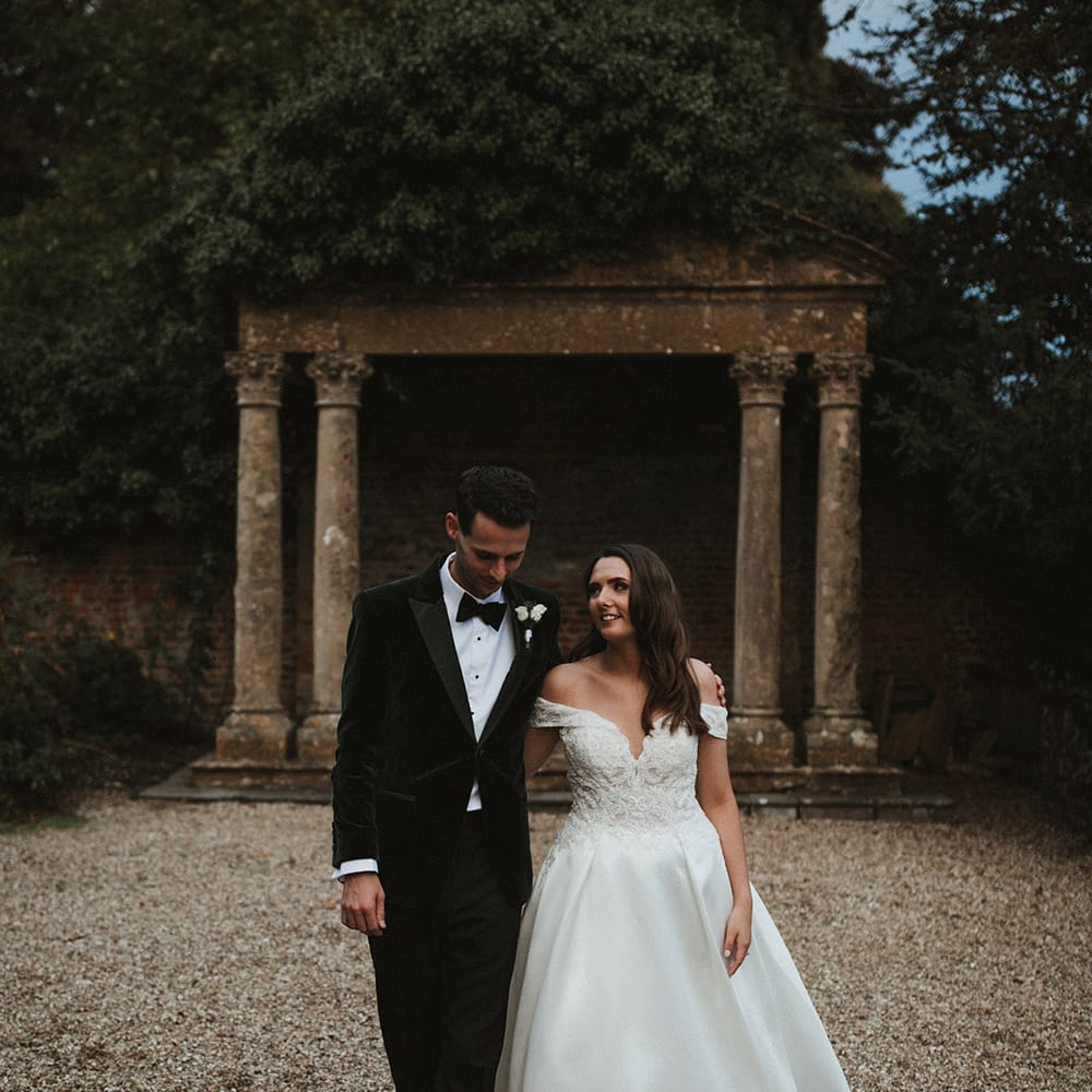 Bride in Pronovias wedding dress with the groom in a velvet tuxedo walking together for their couple portraits at Brympton House 