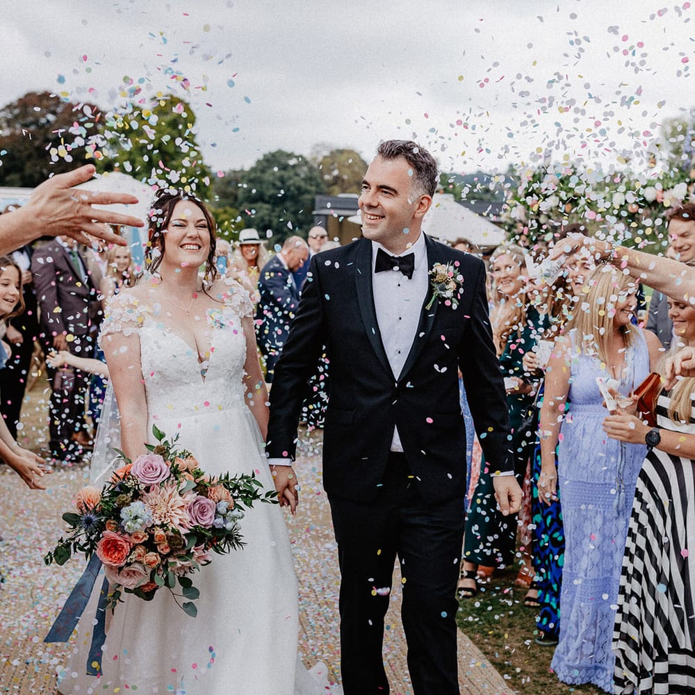 Groom in black tie walks hand in hand with the bride in a flower wedding dress as the guests throw colourful pastel confetti over the couple 