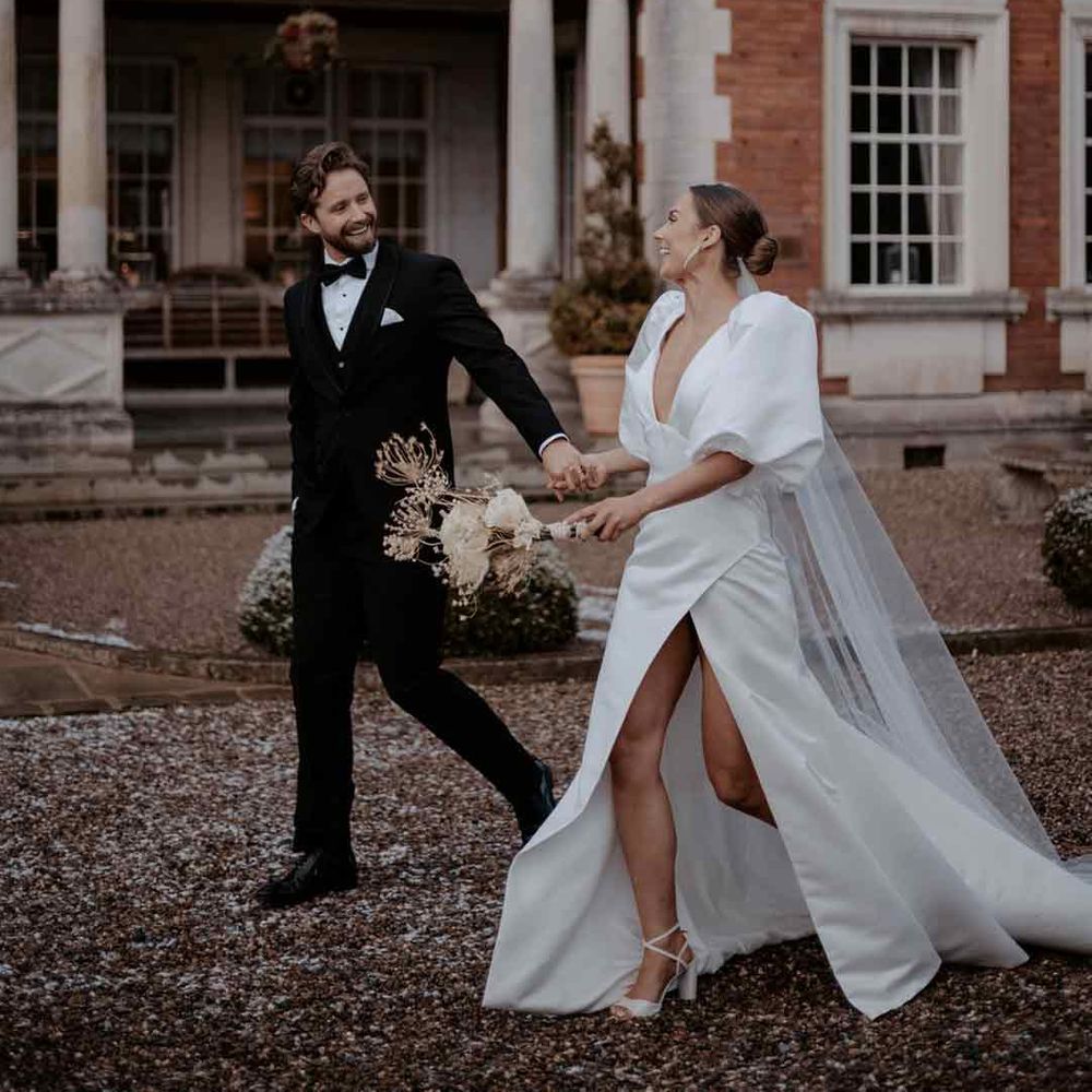 Bride in puff sleeve leg slit wedding dress with church length veil walking with groom in classic black tuxedo - wedding budget spreadsheet 