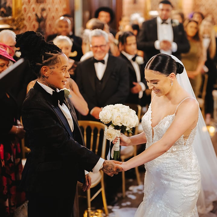 Bride holding white rose bridal bouquet wearing sleeveless v neck Enzoani wedding dress with tulle finish and church length veil holding hands with bride in black snakeskin print tuxedo with black bowtie and white rose boutonniere at the alter of Grittleton House