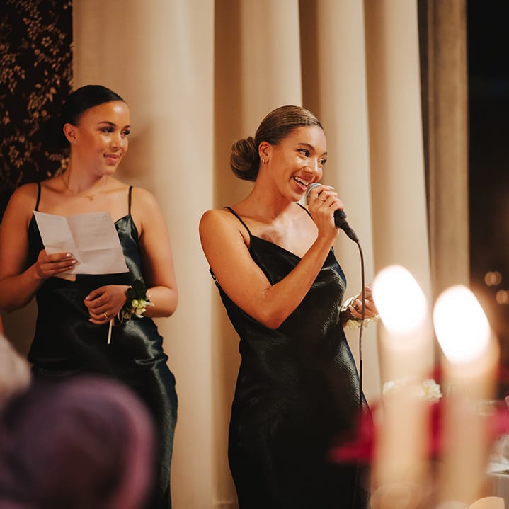 Bridesmaids in strappy satin midi length black bridesmaid dresses doing wedding speeches at Grittleton House wedding venue 