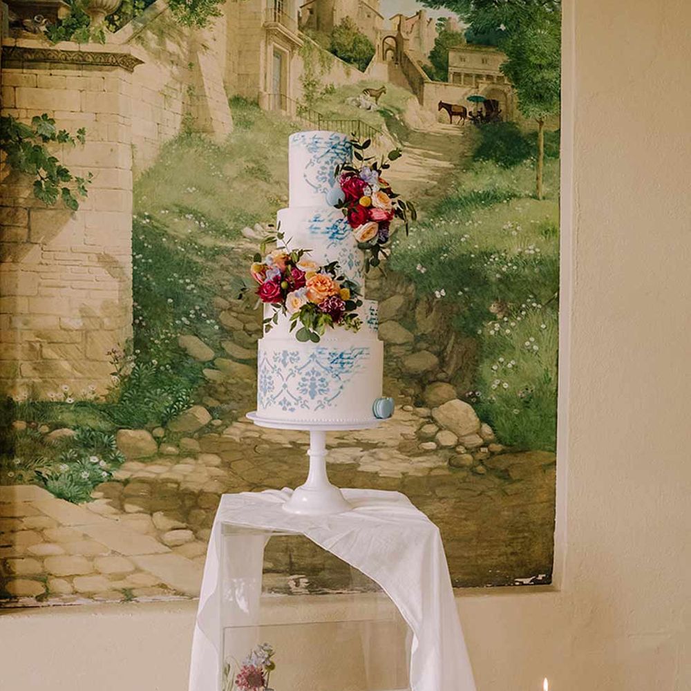 Four tiered toile wedding cake in blue and white on white cake stand on clear glass stand with white wedding drapes and bright blue glass bottles with dried flowers