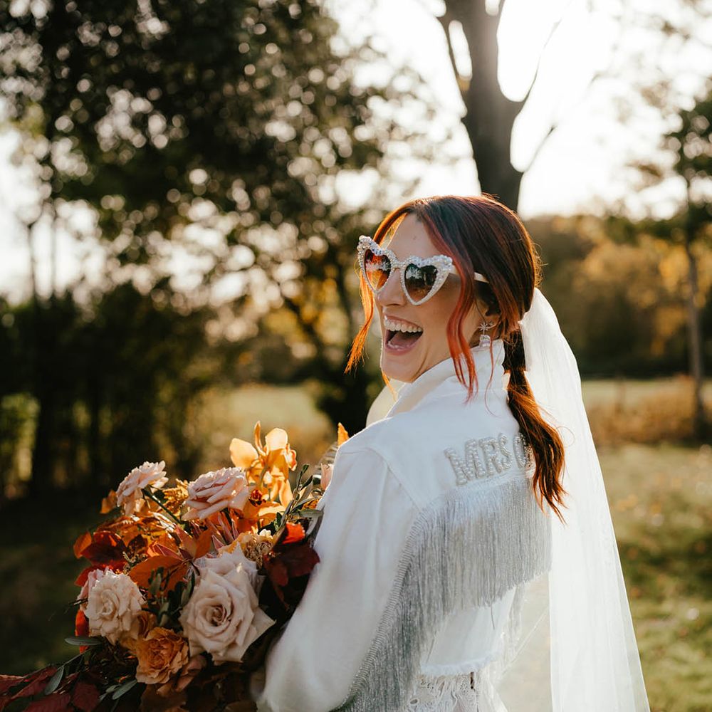Bride in white denim jacket with tassels and pearls with pearl white retro sunglasses holding orange and light pink rose autumnal bouquet