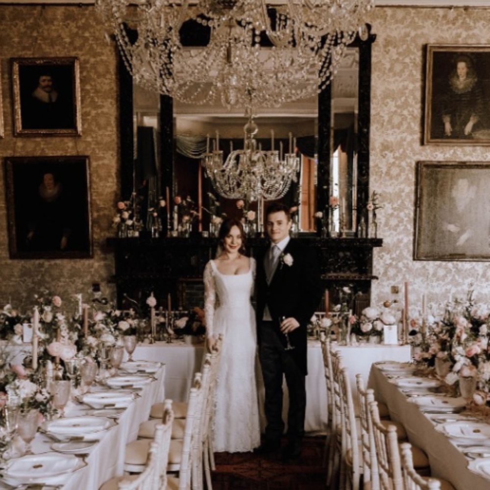 Bride in corset top style long sleeve wedding dress standing with groom in classic back grooms suit in reception room of Cambo House wedding venue with large chandeliers and classic wedding tablescapes 