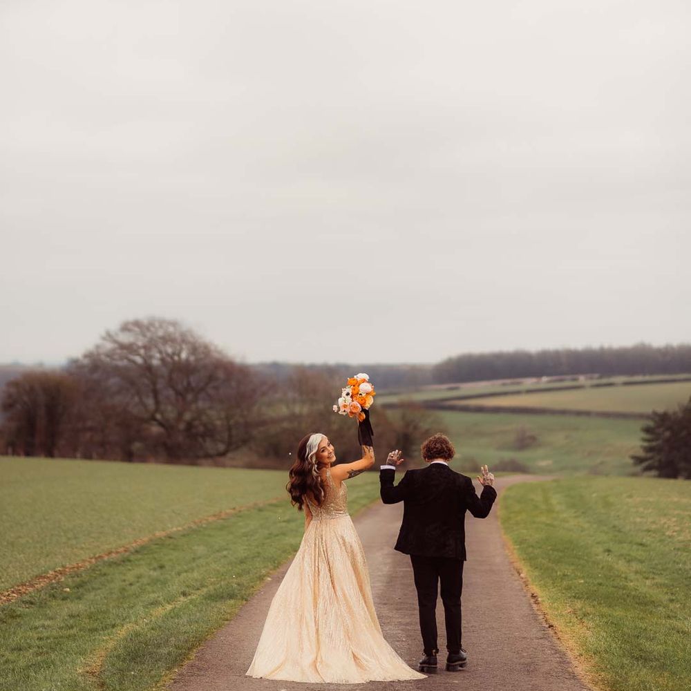 Bride in sleeveless deep v neck sparkly gold wedding dress holding up orange garden rose, white poppy, light pink carnation and foliage bridal bouquet wrapped in sparkly gold ribbon with bride in dark bridal suit at Kingscote Barn Tetbury 