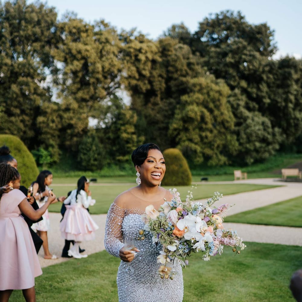 Bride in illusion v neck sheer off shoulder long sleeve pearl embellished wedding gown holding large pastel and foliage mixed bridal bouquet on the grounds of Syon Park