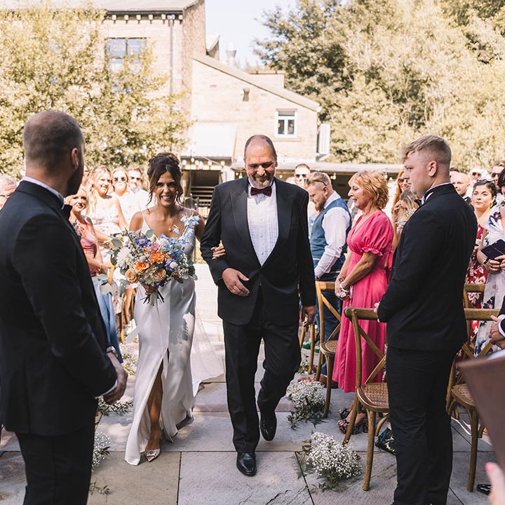 Father of the bride in black tuxedo walks the bride down the aisle at outdoor wedding 