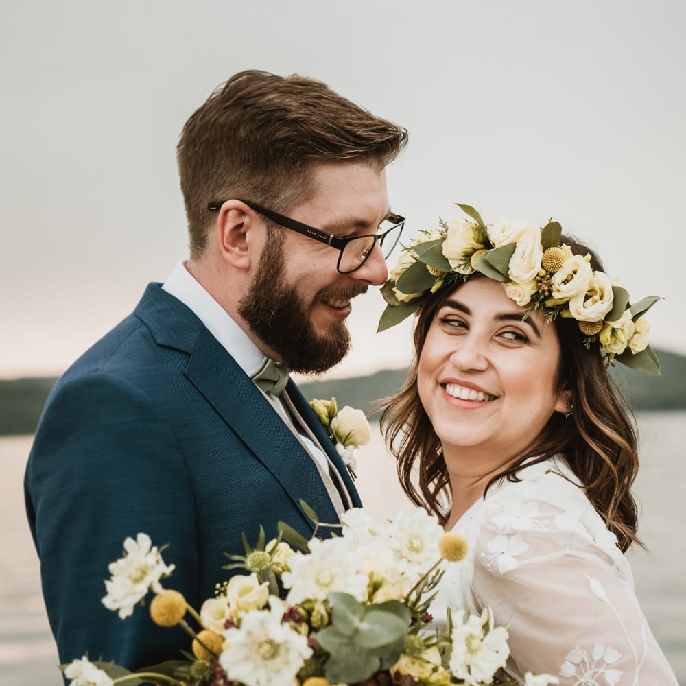 White bearded groom in a navy suit with glasses looking at his boho bride wearing a yellow, white and green flower crown 