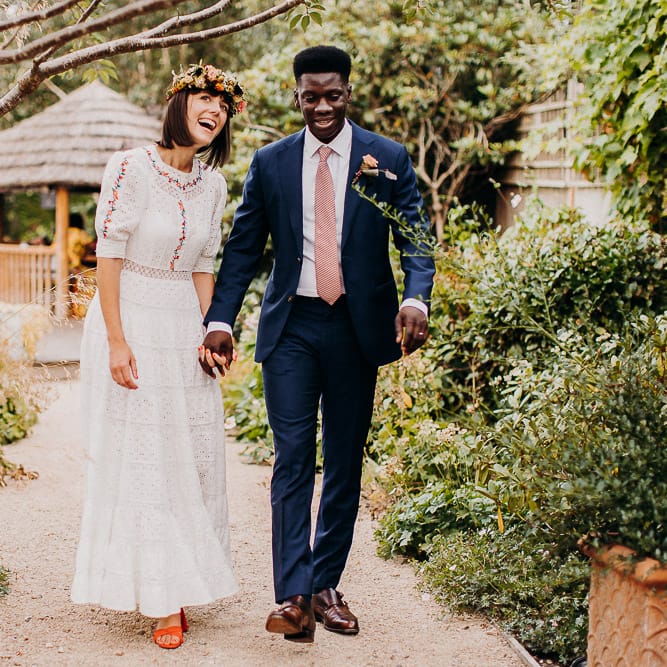 Groom in a navy suit and pink tie holding his brides hand in a white Broderie Anglaise wedding dress and colourful flower crown in a garden 