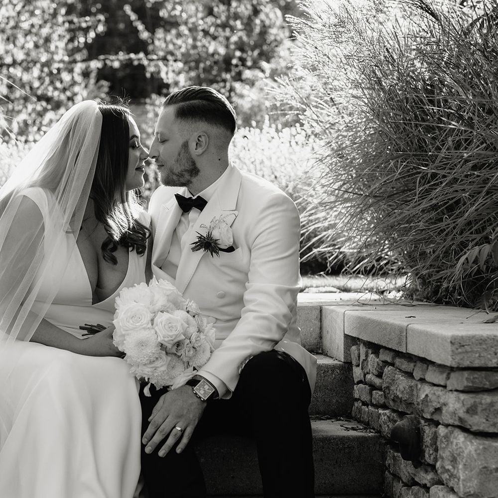 Bride in V neck Made With Love wedding dress and veil holding white rose bouquet sits outside with groom in white double breasted tuxedo jacket
