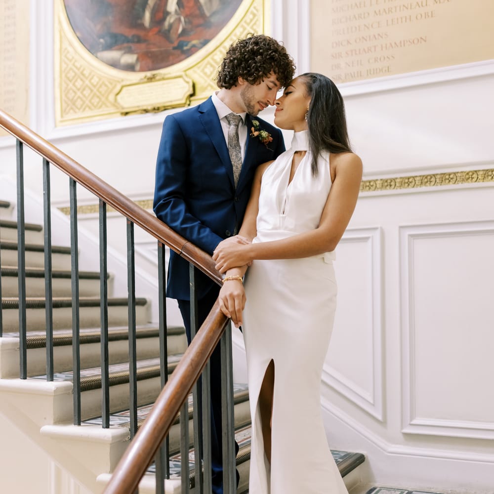 Bride & groom stand on staircase whilst kissing as bride wears high neck gown with split to the front