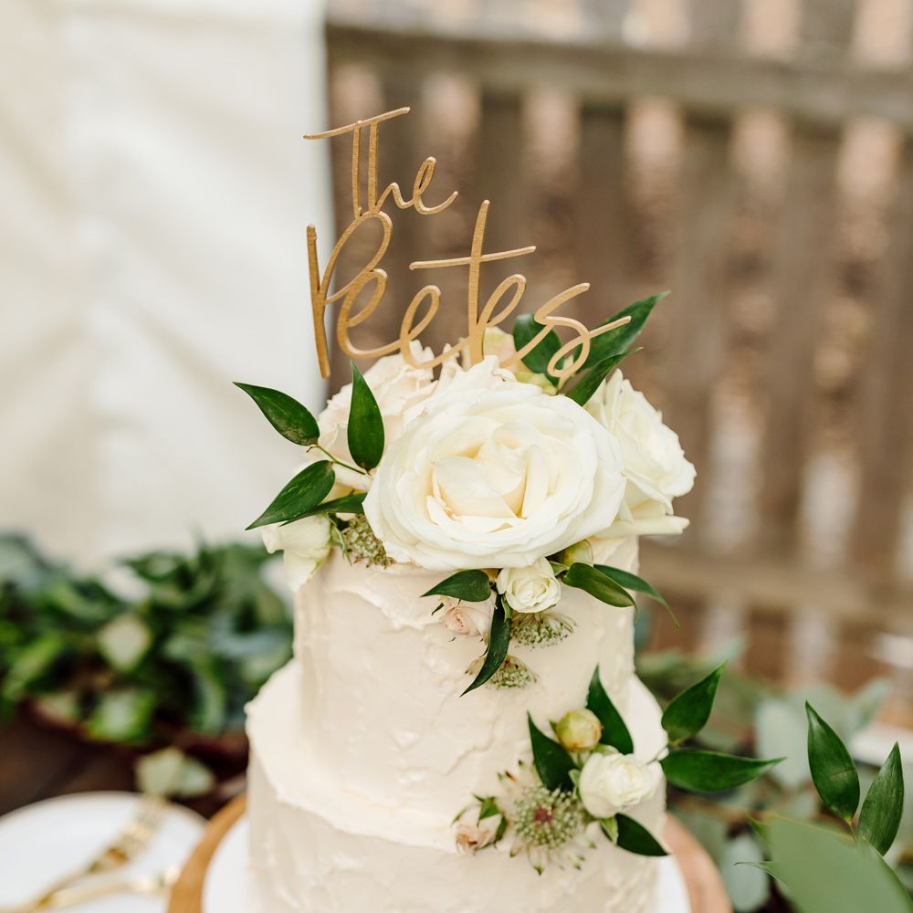 White two tier wedding cake with white rose and green leaf decoration with cake topper reading 'The Peetes' on wooden table at backyard wedding
