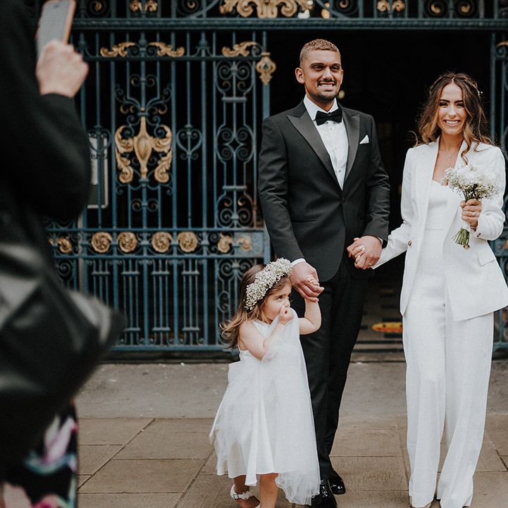 Family portrait with a groom in a tuxedo, bride in a suit and flower girl wearing a gypsophila flower crown 