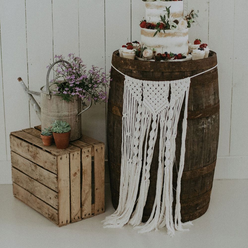 White three tier semi naked wedding cake with berry and foliage decor on wooden barrel with macrame hanging next to rustic wedding decor at Isle of Wight wedding
