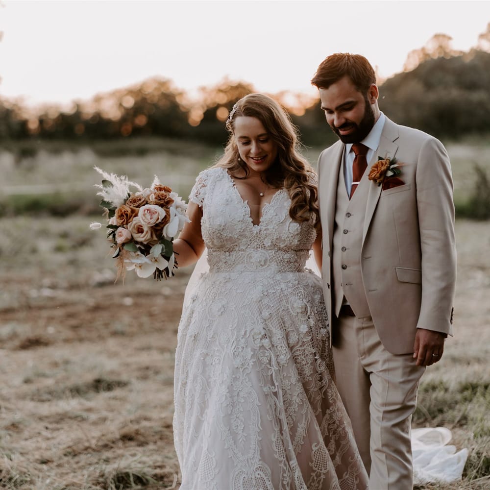 Birde in homemade lace wedding dress with train holding mixed rose bridal bouquet walks through field with groom in three piece beige suit, burgundy tie and floral buttonhole after DIY garden wedding ceremony