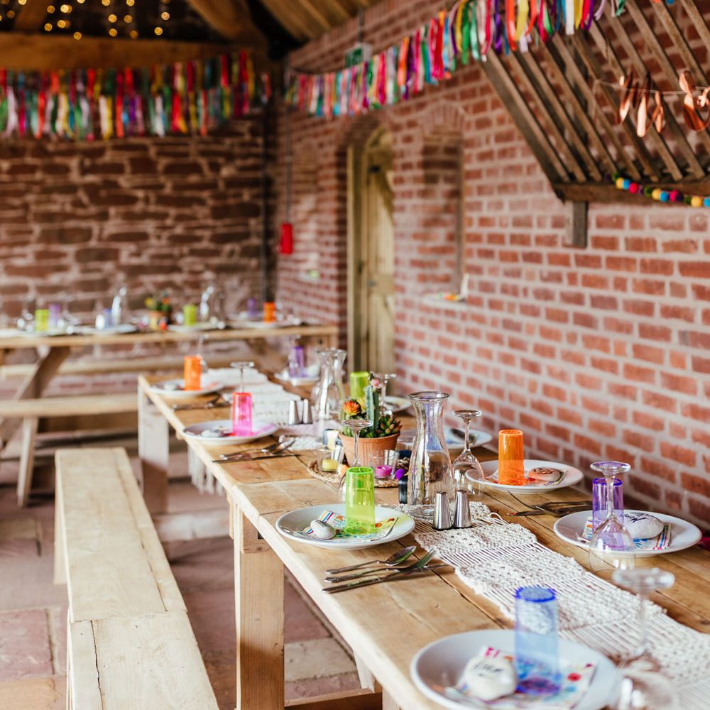 Long wooden wedding table with benches, colourful cups, macrame table runner and colourful streamers for rustic wedding breakfast at The Bridal Barn in Claverley