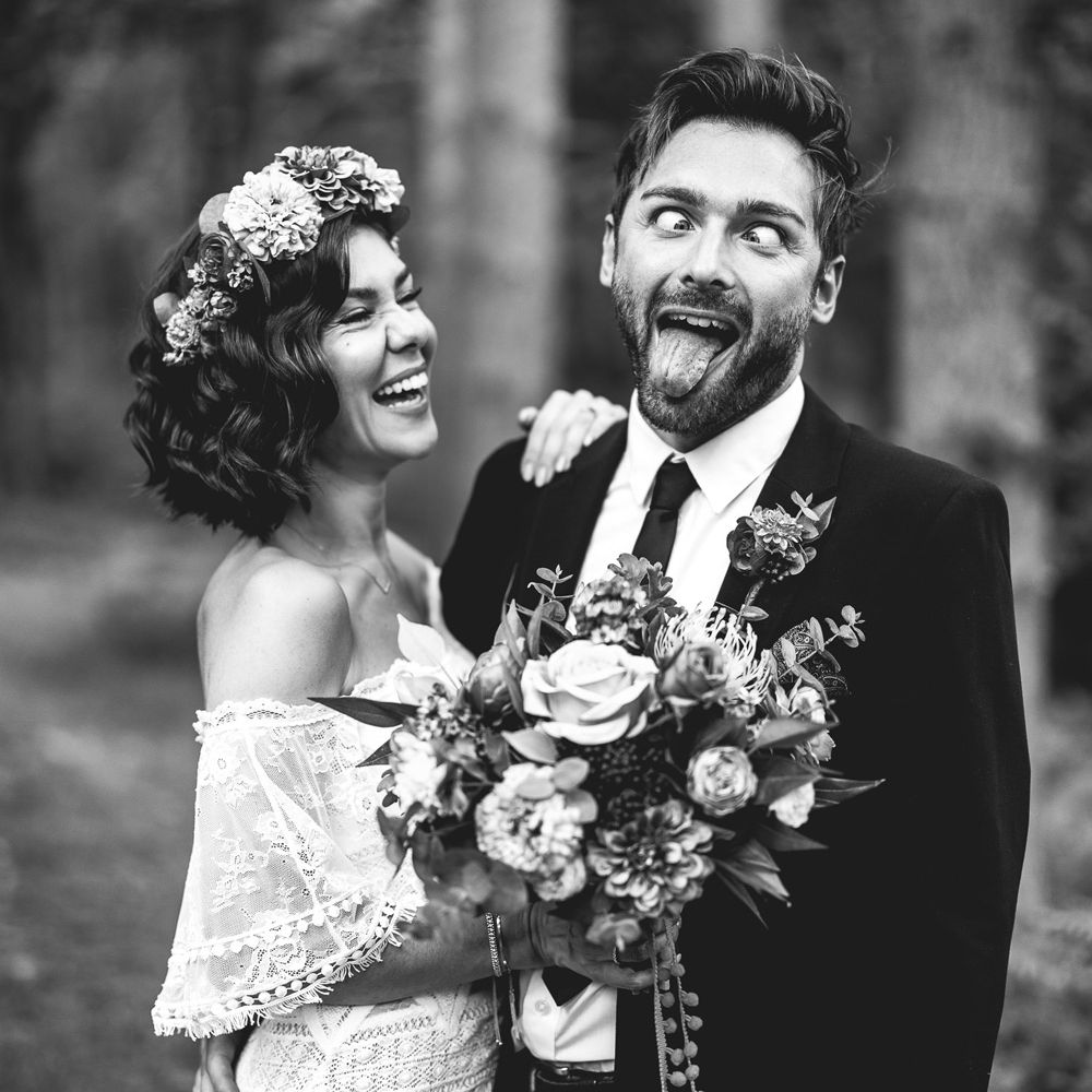 Bride in off the shoulder Grace Loves Lace wedding dress and colourful flower crown holds colourful wedding bouquet as she stand with groom in black suit as he pulls a face 