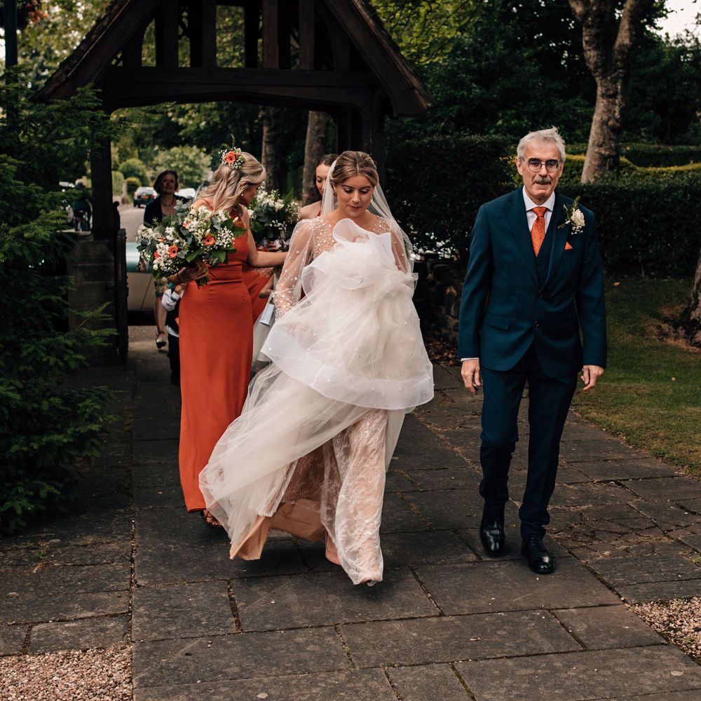 Bride in Julie Vino wedding dress and veil holds skirt as she walks to the church with father in blue three piece suit and bridesmaids in orange dresses before wedding ceremony