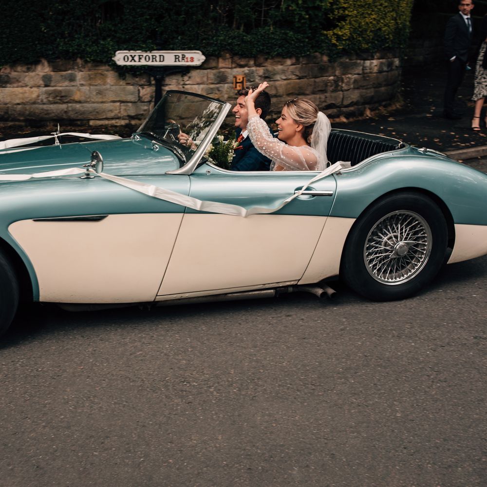 Bride in long mesh sleeve wedding dress and veil waves as groom in blue checked suit drives white and blue vintage sports car from church ceremony