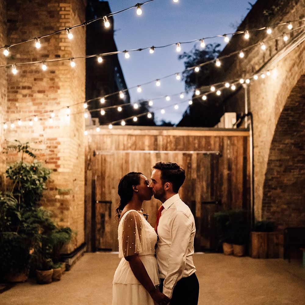 Black bride and groom kissing under a canopy of festoon lights at their 100 Barrington wedding 
