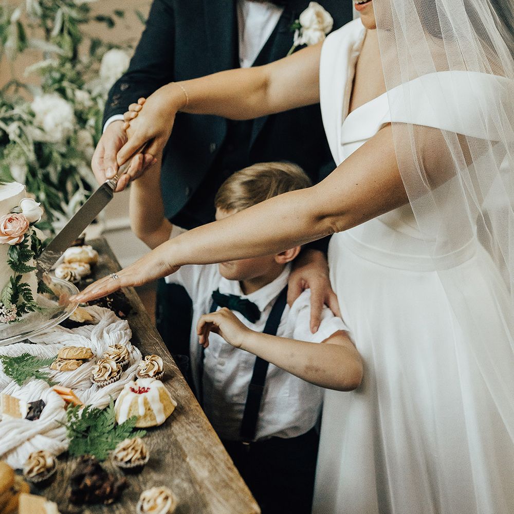 Bride in an off the shoulder wedding dress and veil cutting the cake with there groom and son 