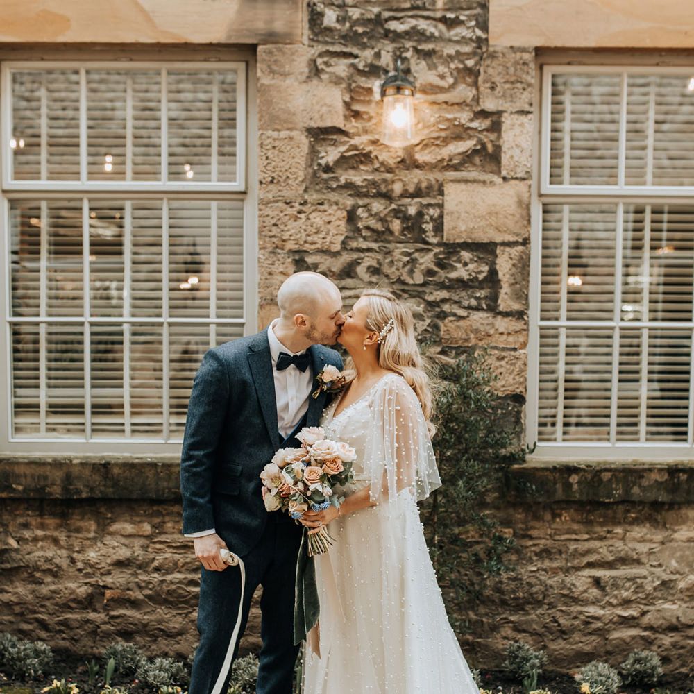 Bride in white pearl encrusted wedding dress with sleeves and train kisses groom in navy tweed suit with bow tie holding French bulldog on a lead