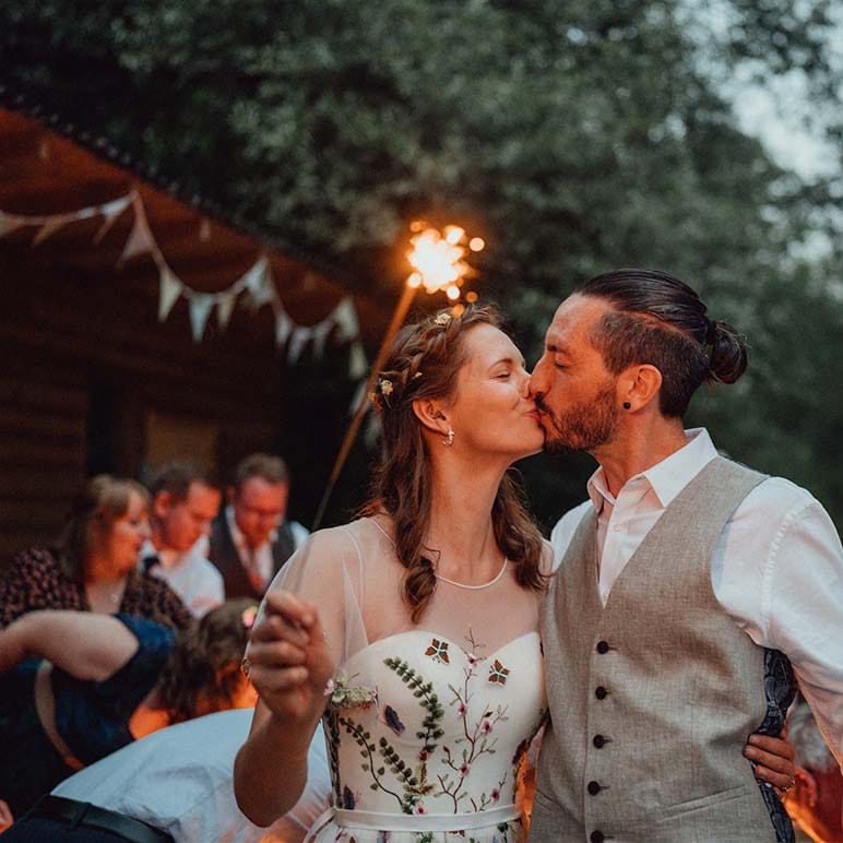 Groom in white shirt and grey waistcoat hugs with bride as they hold sparklers 