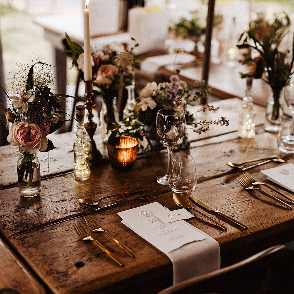 Wooden banquet tables with gold candle holders and cutlery with white menu card with wax seal with pink and white wedding flower arrangement