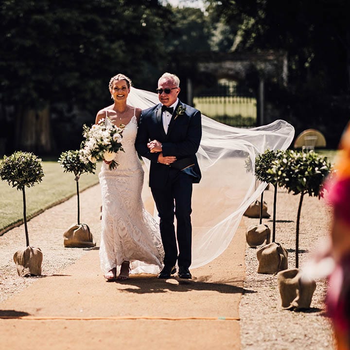 Father of the bride in blue tuxedo walks the bride down the aisle in lace wedding dress 