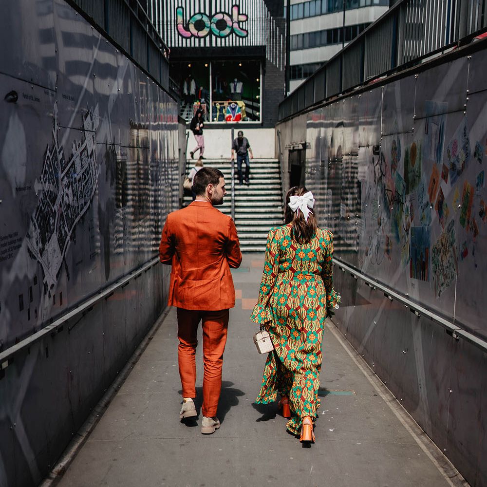 Bride wearing white bow in her brown hair and carrying nude purse walk along street in Bristol with her groom wearing orange suit 