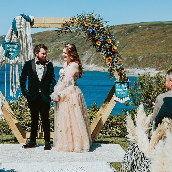 Bride & groom stand beneath DIY wooden arch finished with florals and wedding banners during ceremony outdoors on beach front in Cornwall