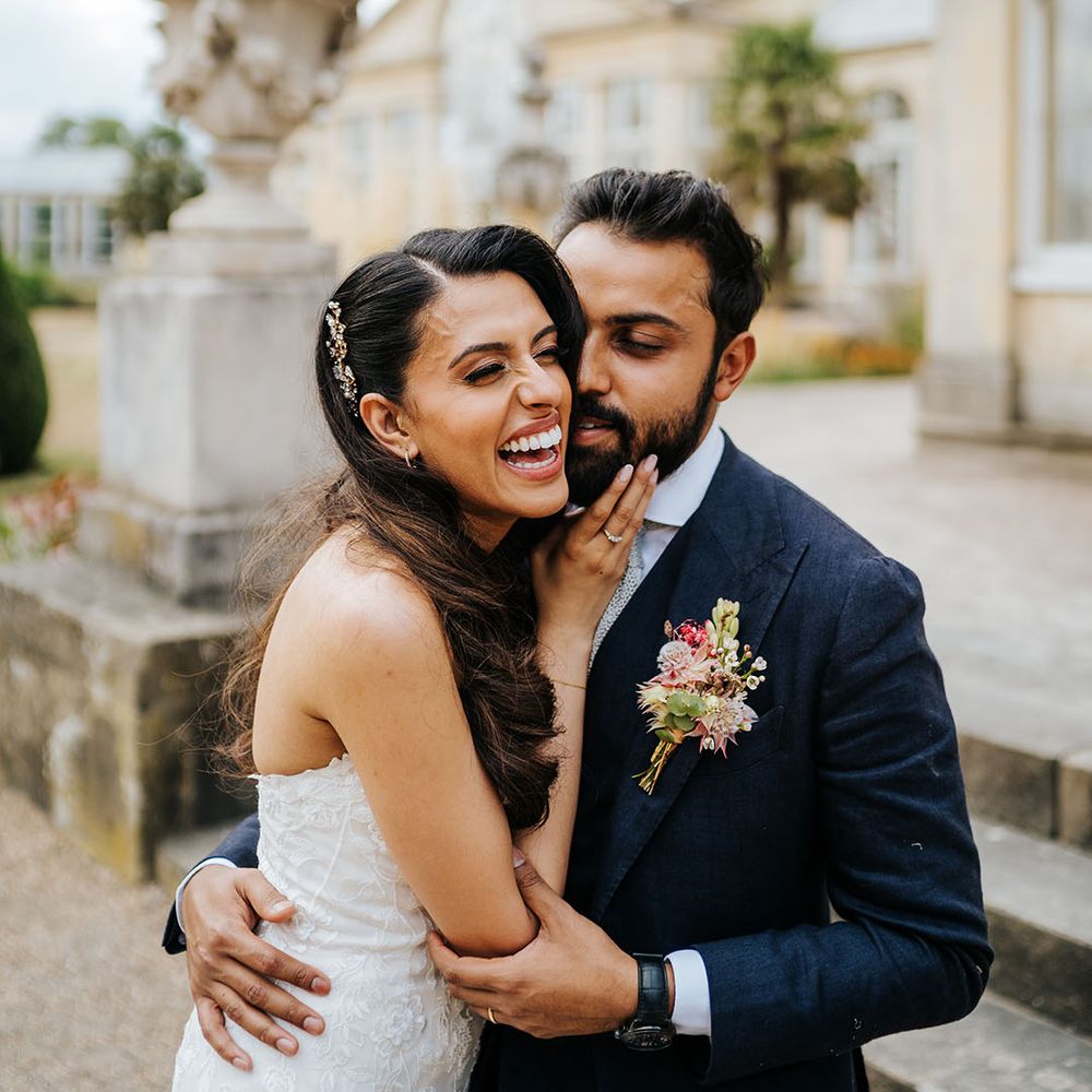 Groom wears three piece blue suit with floral buttonhole and stands beside his bride outdoors at Syon Park