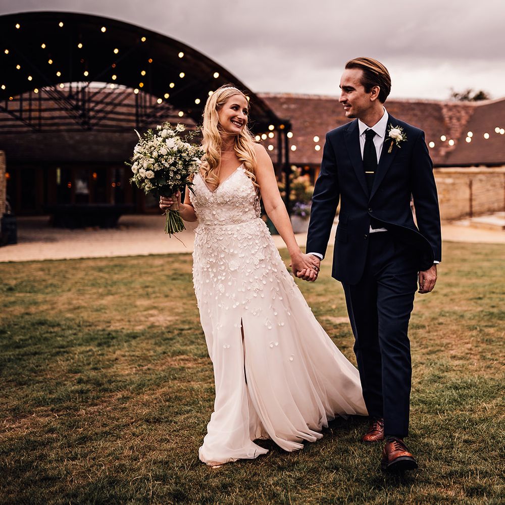 Bride and groom walk around outside their barn wedding venue holding hands together 