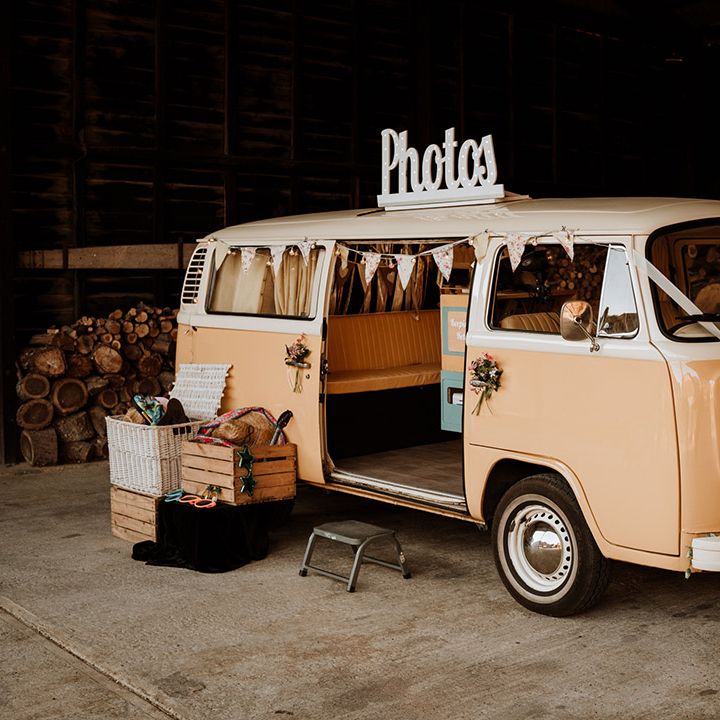 A pink VW camper photo booth decorated with white ribbon and sunflowers with a box of props for photos 