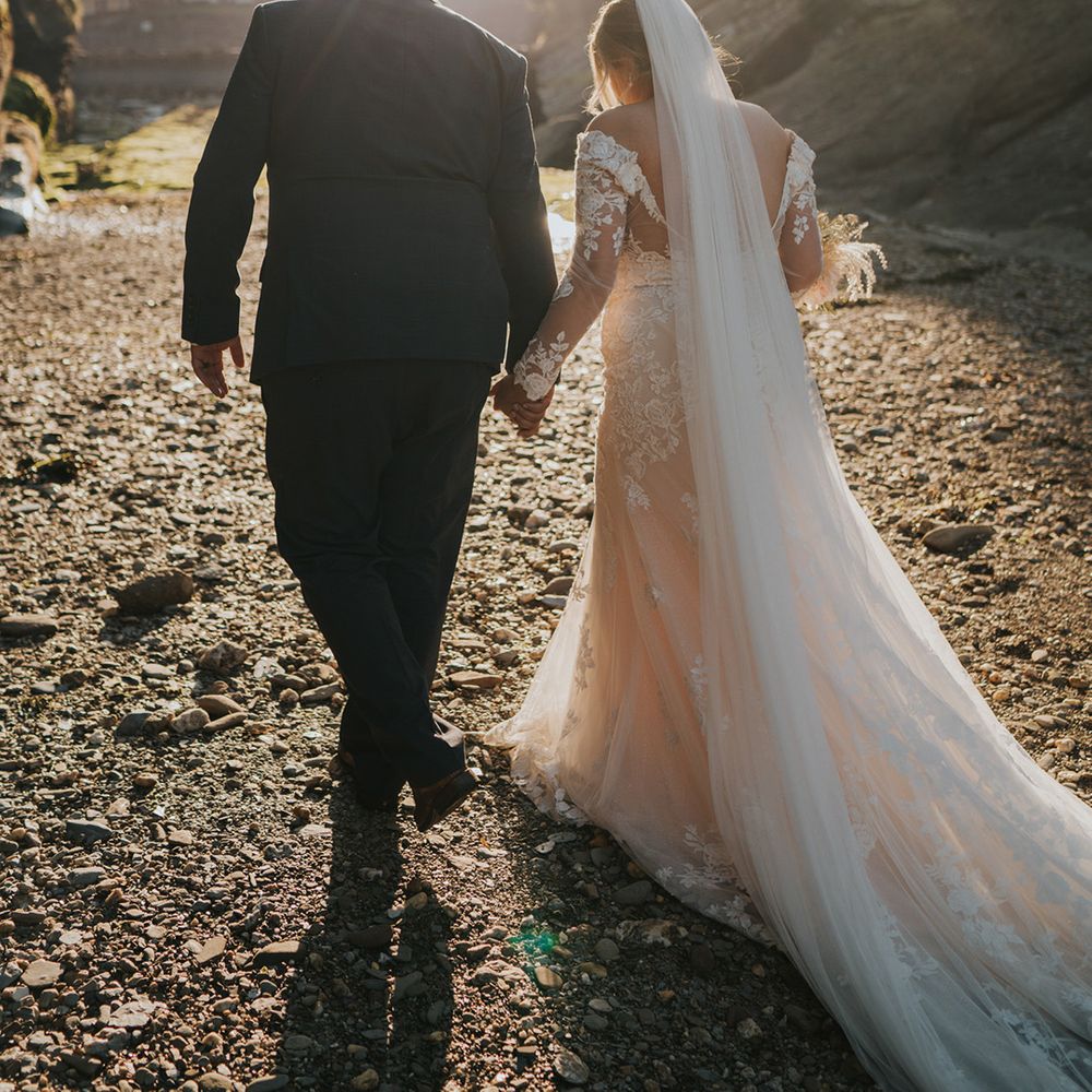 Bride walks along the beach as the sun begins to set with her groom