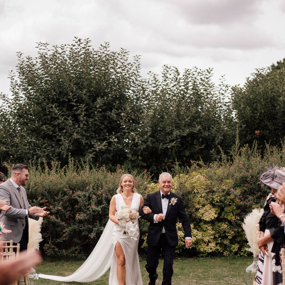 Father of the bride in black tie walks the bride in a plunging white wedding dress down the aisle 