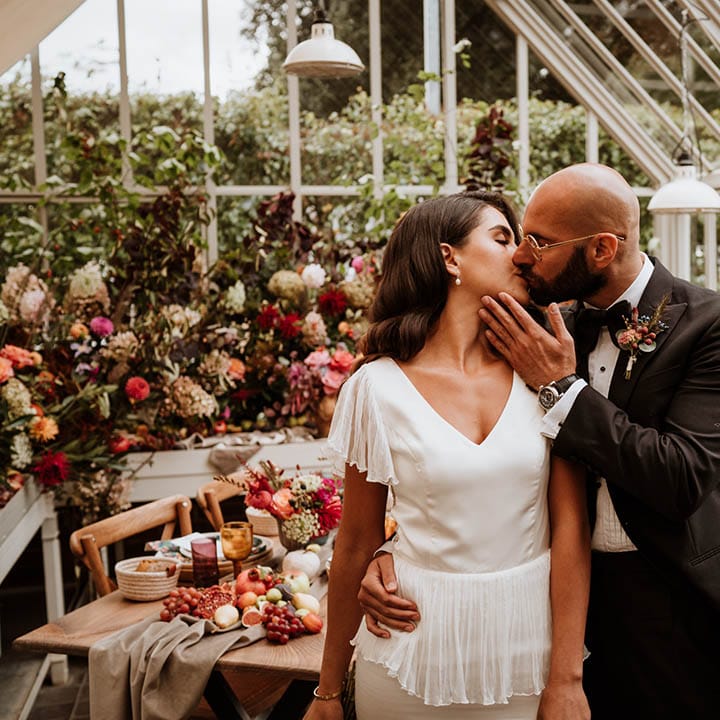 Groom in classic black tuxedo with autumnal dried flower boutonniere and black bowtie kissing bride in v neck satin wedding dress with sheer butterfly sleeves in glasshouse wedding venue