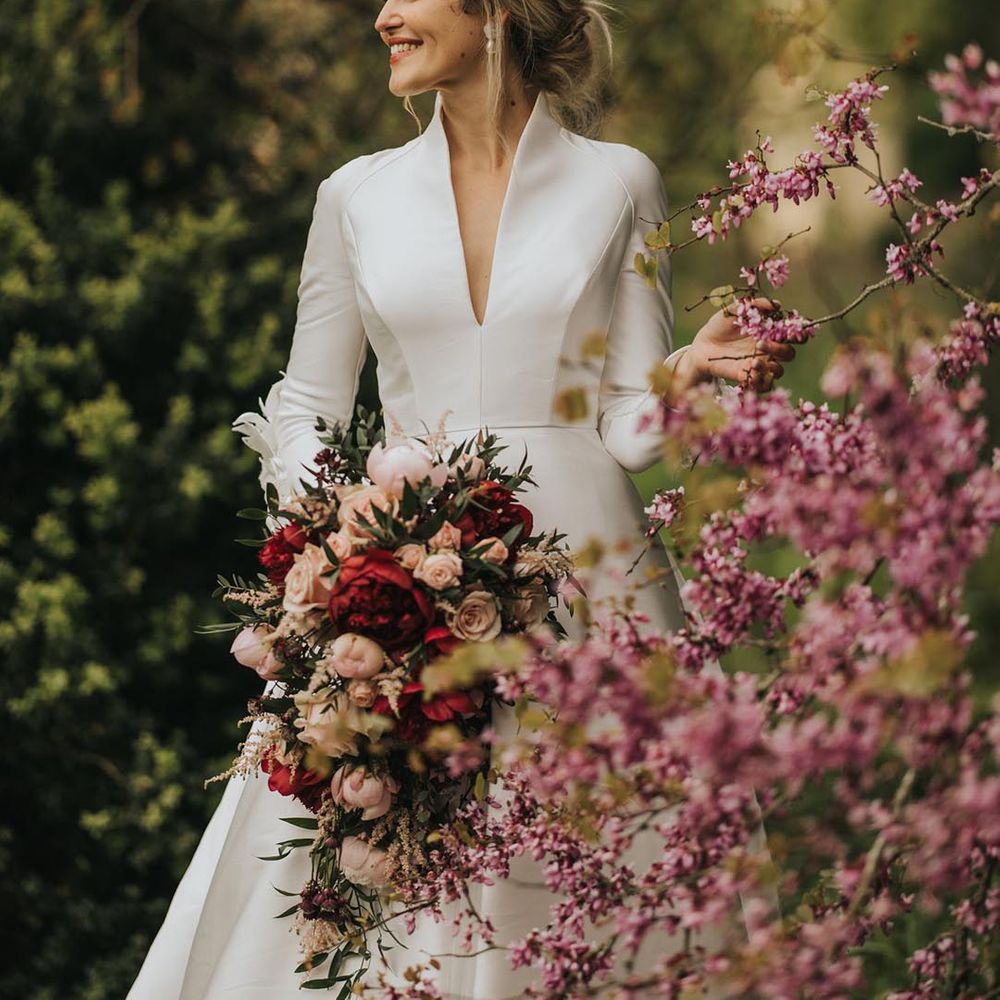Bride in Jesus Peiro high neck long sleeve v neck wedding dress with pearl earrings, a floral hair piece and red and pink bouquet