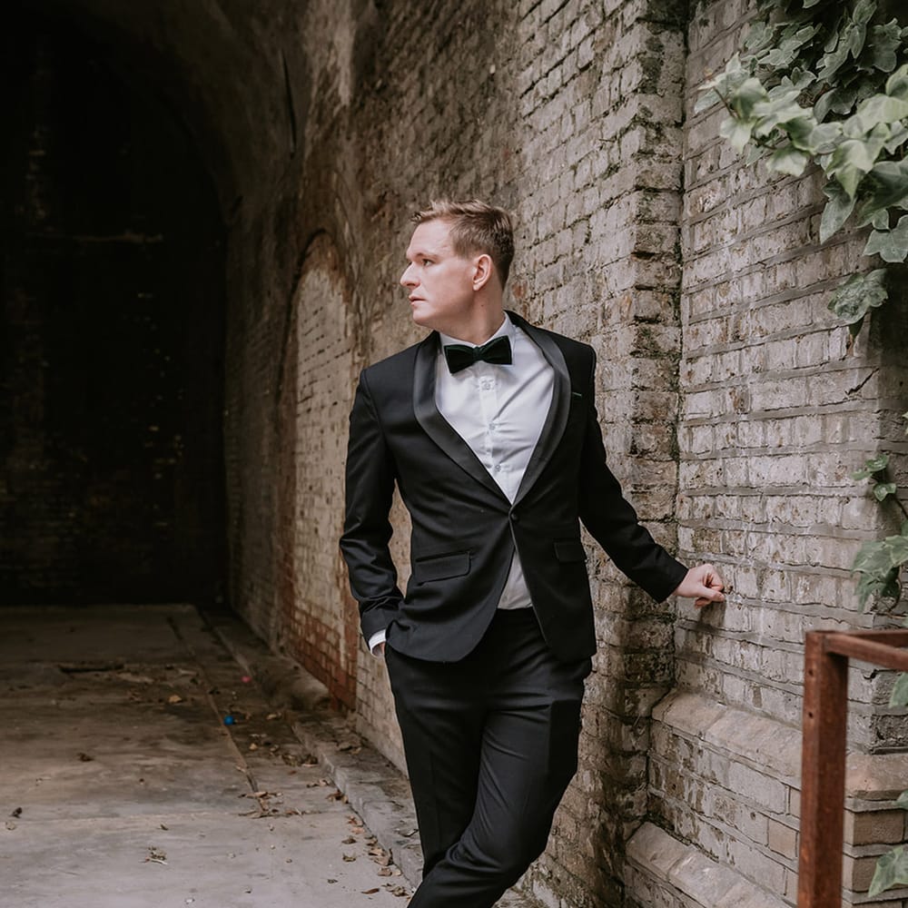Groom in stylish black tuxedo with black bowtie with light orange rose boutonniere at multicultural Christmas wedding with Lion Dance
