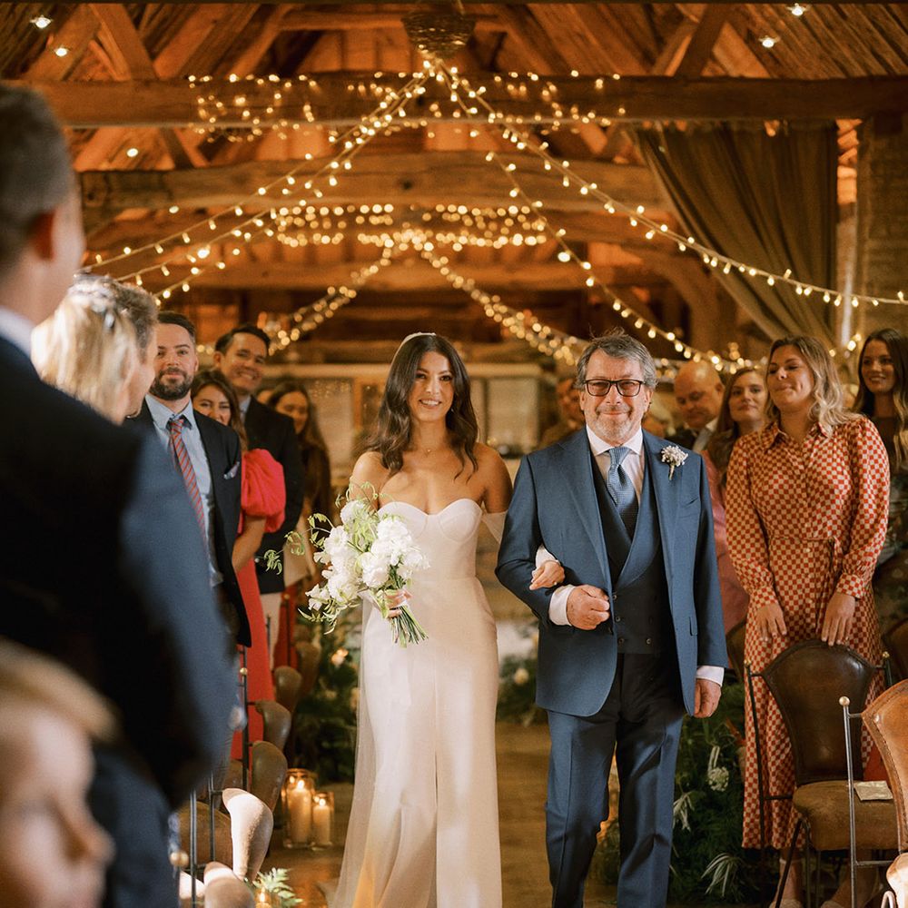 Father of the bride in a navy blue suit walking the bride down the aisle in a strapless wedding dress with detachable pearl sleeves for rustic barn wedding