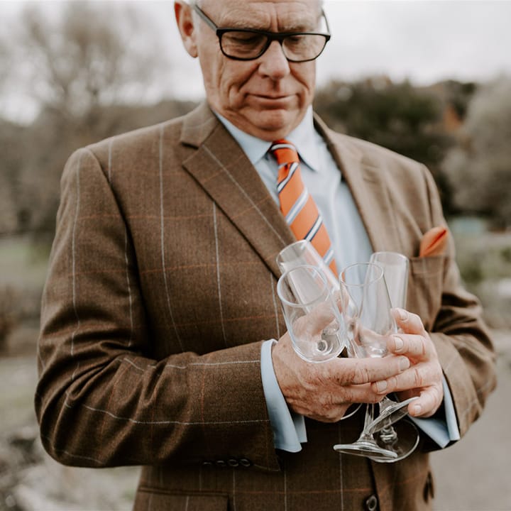 Wedding guest in brown suit with orange checkered detail for autumnal wedding outfit holding champagne glasses 