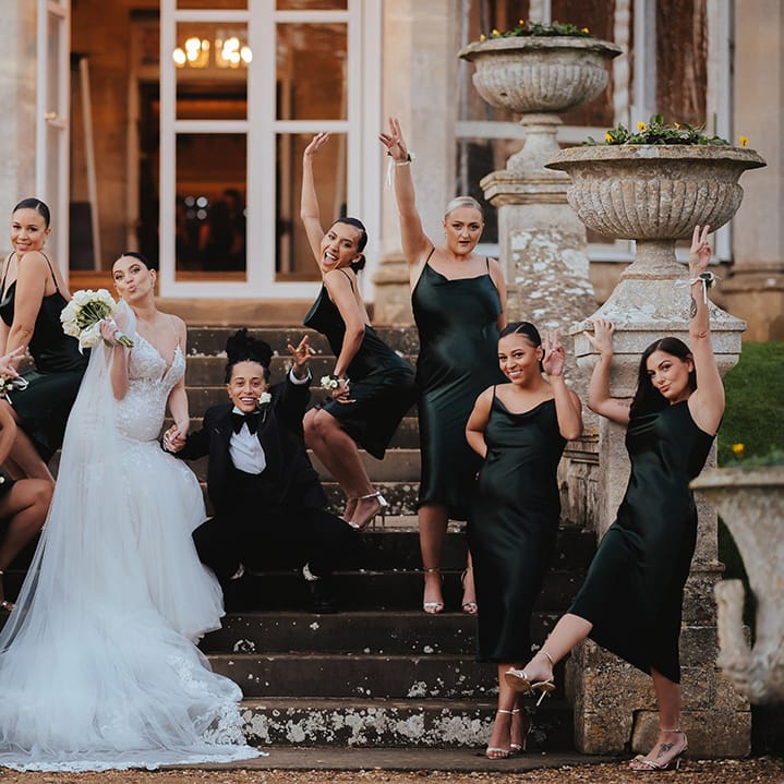 Bridesmaids in midi length black bridesmaid dresses posing with brides in lace sleeveless v neck Enzoani wedding dress with tulle finish and church length veil and black snakeskin print tuxedo with black bowtie and white rose boutonniere at Grittleton House
