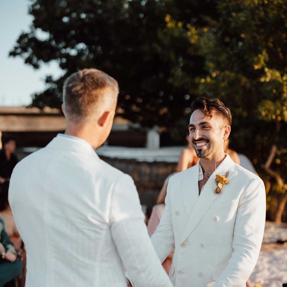 Groom in mens white wedding suit, dusky orange garden rose and dried flower boutonniere, white shirt, and silver jewellery holding hands with groom in white double breasted suit 