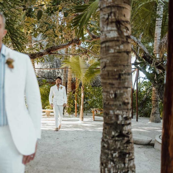  Groom wearing white blazer, light blue and white striped shirt, dusky orange garden rose and dried flower boutonniere, white suit trousers and gold jewellery doing first look photo with groom in white blazer with dusky orange garden rose and dried flower boutonniere, white shirt, white suit trousers 