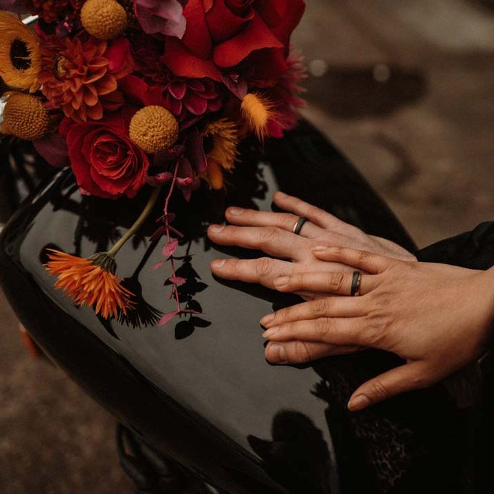 Bride and groom wearing black wedding rings on the back of a motorbike with red vibrant flower arrangements