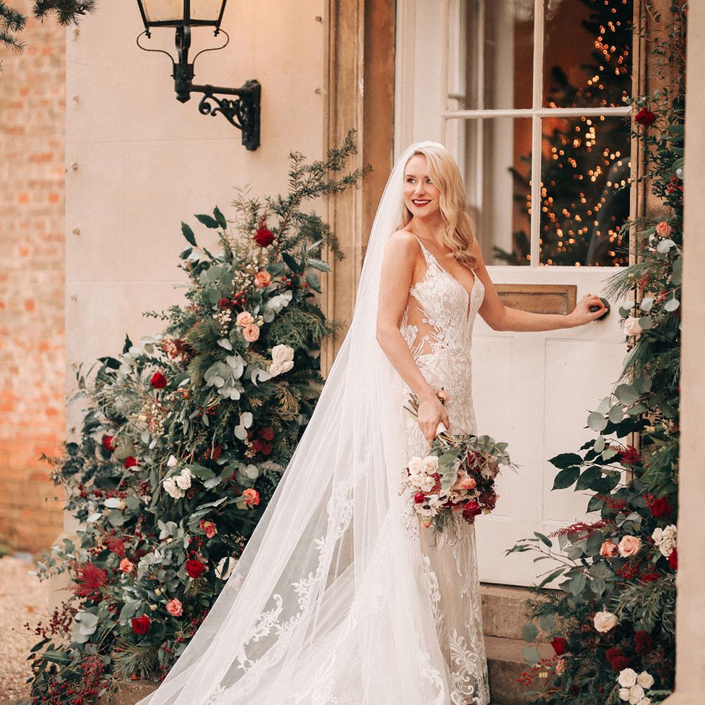 Bride in embellished wedding dress with delicate embroidered veil walking into venue with Christmas tree decor and flower columns 