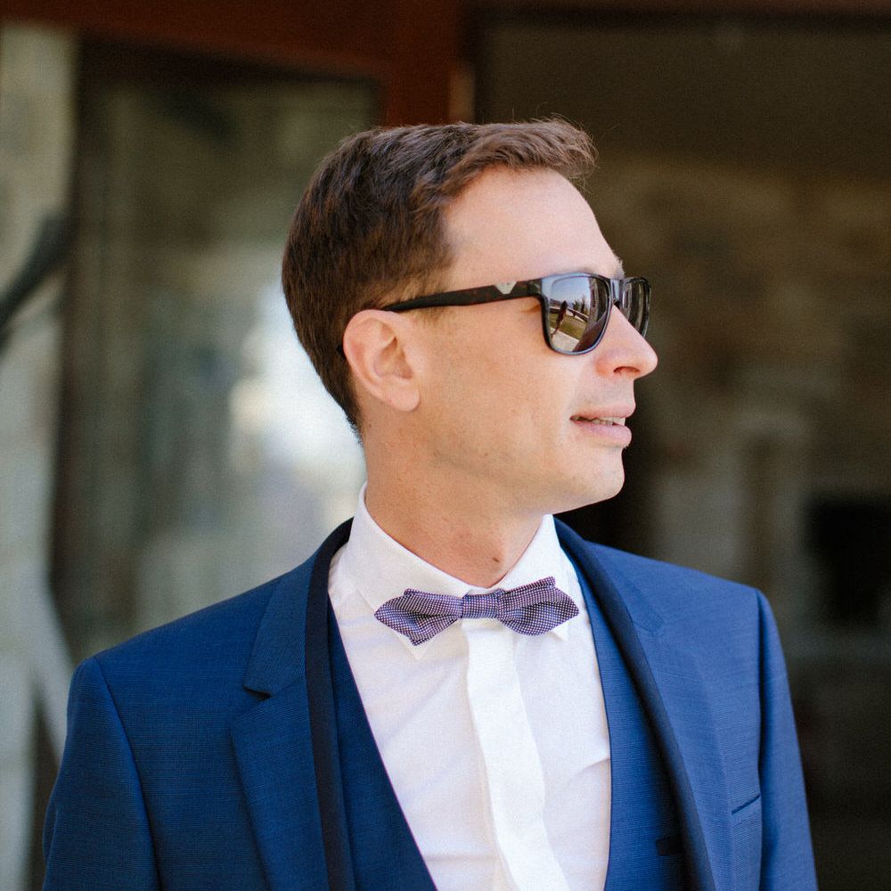 The groom wearing a dark blue three piece suit with a bow tie and sunglasses