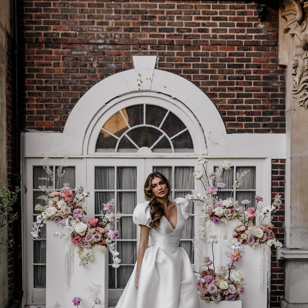 Bride in a stylish Love In Lace Bridal gown with puff sleeves and front split standing next to blush pink floral arrangements 